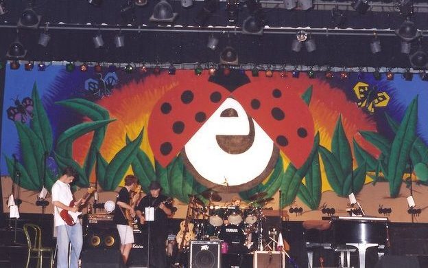 The theatre concert backdrop features a giant red ladybug in front of a brightly colored sunset with oversized grass, ants and butterflies that are so large the humans on stage look tiny. The ladybug wings open to display the initial "e" for "expressions," the name of the concert. Wings are open in this picture and a band is warming up on stage.